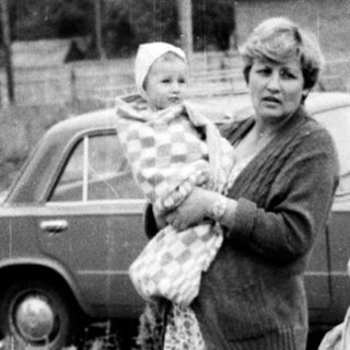 Three women standing outdoors with a baby, holding groceries near vintage cars.