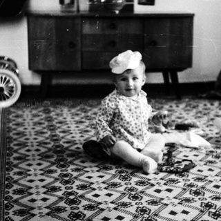 Baby wearing hat sitting on patterned rug indoors.