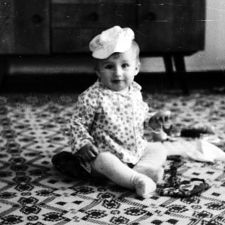 Baby wearing hat sitting on patterned rug indoors.