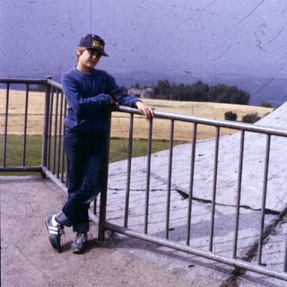 Boy in cap and blue tracksuit posing with skateboard on elevated platform.