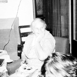 Old woman sitting indoors with family during television broadcast.