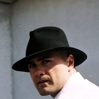 Man in white shirt and fedora near real estate sign.
