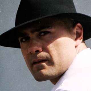 Man in white shirt and fedora near real estate sign.