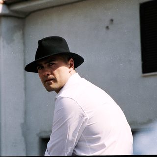 Man in white shirt and fedora near real estate sign.