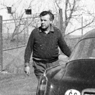 Vintage car with Czech license plate, man standing beside it near rural house.