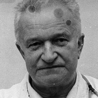Older man in medical coat with small scars on forehead.
