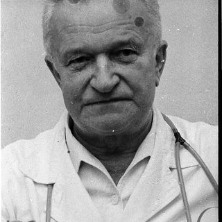 Older man in medical coat with small scars on forehead.