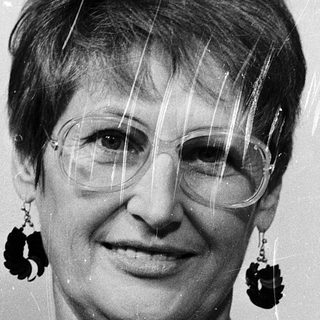Woman wearing glasses, patterned blouse, dangly earrings.