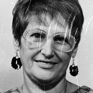 Woman wearing glasses, patterned blouse, dangly earrings.