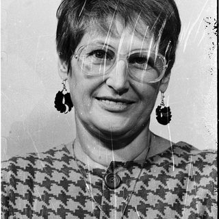 Woman wearing glasses, patterned blouse, dangly earrings.