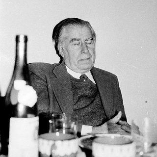 Elderly man seated at a table with wine bottle.