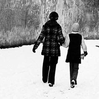 Two children walking in snow beside a wall.