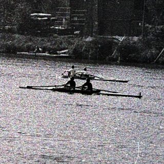 Rowing team on calm river near shoreline buildings.