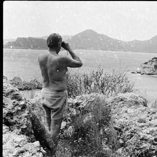 Shirtless man taking photo of rocky island in snowy coastal landscape.