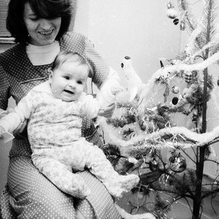 A woman holds a baby near a decorated Christmas tree.
