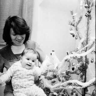 A woman holds a baby near a decorated Christmas tree.