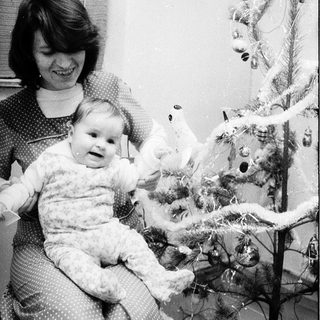 A woman holds a baby near a decorated Christmas tree.