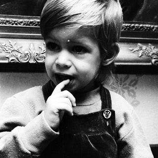 Young child in overalls, touching finger to lip.