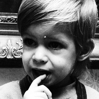 Young child in overalls, touching finger to lip.