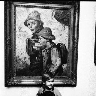 Child posing beside vintage portrait of woman in outdoor attire.