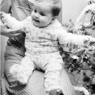 A woman holding a smiling baby indoors near holiday decorations.