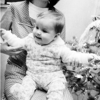 A woman holding a smiling baby indoors near holiday decorations.