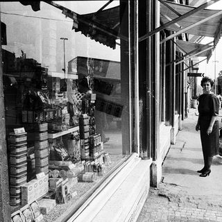 Small street shop with canned goods displayed, two women walking outside.