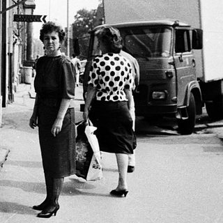 Small street shop with canned goods displayed, two women walking outside.