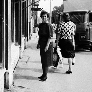 Small street shop with canned goods displayed, two women walking outside.
