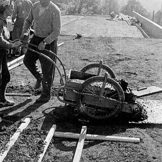 Construction workers using manual drilling equipment on pavement.