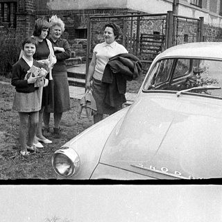 Four women standing by vintage car in snowy residential area.