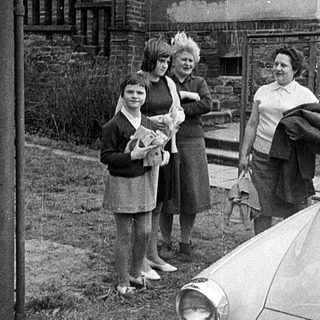 Four women standing by vintage car in snowy residential area.