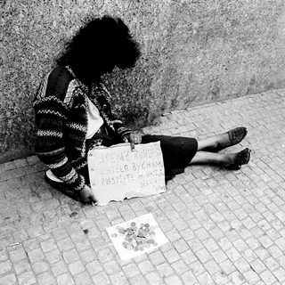 Person sitting on pavement holding sign requesting help.