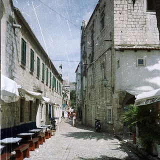 Narrow cobblestone street with historic buildings, outdoor seating, light rain.