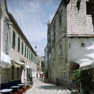 Narrow cobblestone street with historic buildings, outdoor seating, light rain.