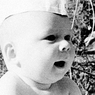 Baby wearing crown sitting outdoors on blanket.