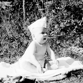 Baby wearing crown sitting outdoors on blanket.