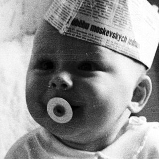 Baby wearing paper hat, sucking pacifier indoors.
