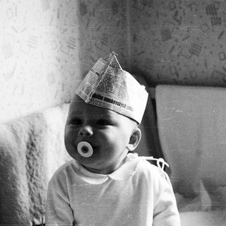 Baby wearing paper hat, sucking pacifier indoors.
