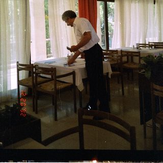 Man setting table indoors near large windows.