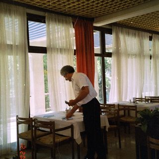 Man setting table indoors near large windows.