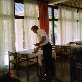 Man setting table indoors near large windows.