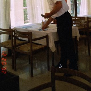 Man setting table indoors near large windows.