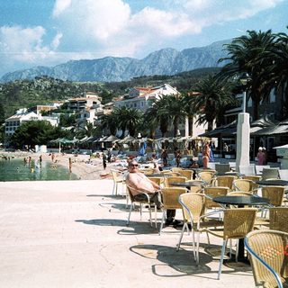Sunny coastal beachfront with palm trees, seating area, and mountain backdrop.