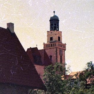 Old stone building with red roof, church tower in background.