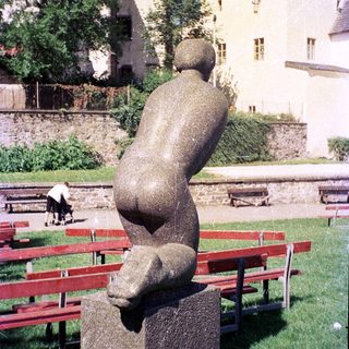 Abstract sculpture of seated figure in park setting.