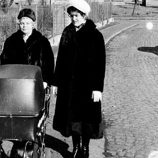 Two women in winter clothing push a baby carriage on a residential street.