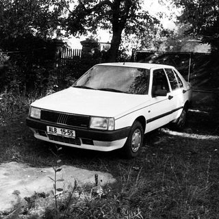 White sedan parked in overgrown driveway.