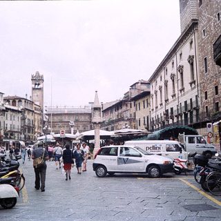 Historic European city street with pedestrians, scooters, and market stalls.
