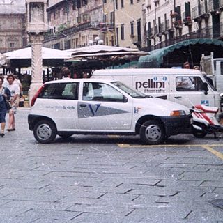 Historic European city street with pedestrians, scooters, and market stalls.